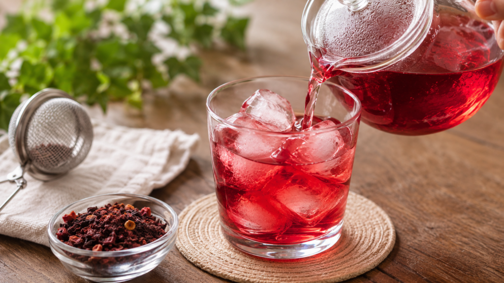 A realistic close-up of deep red berry herbal tea poured over large ice cubes in a glass, with a glass teapot, loose fruit tea blend, and tea infuser on a wooden table, illustrating the on-the-rock brewing method.