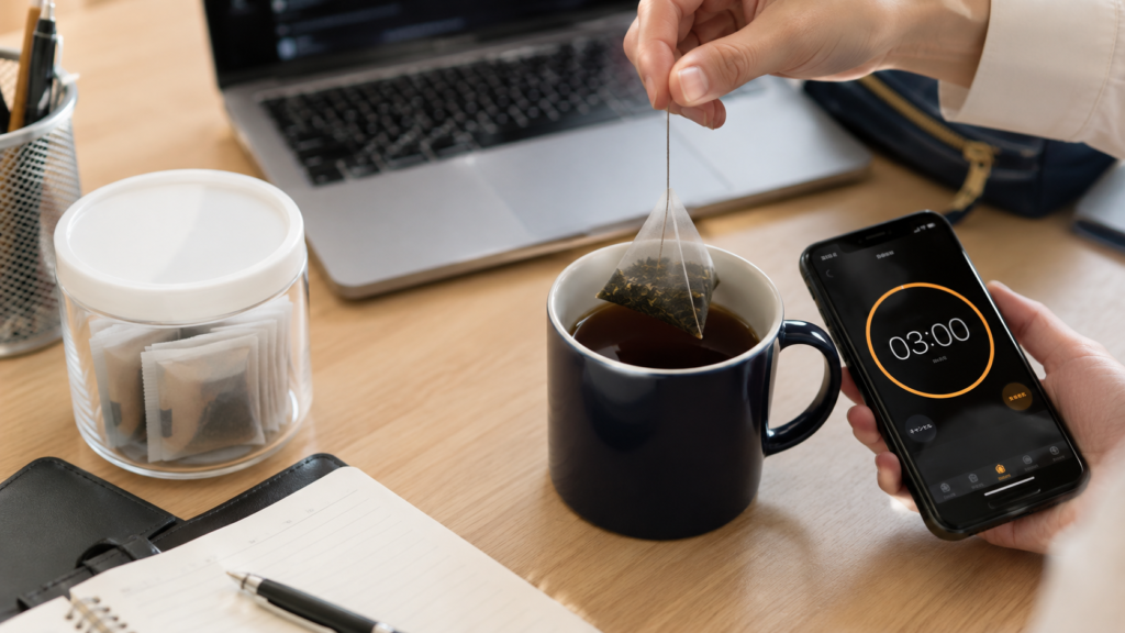 A realistic office desk with a mug of tea, a pyramid tea bag, a smartphone set to a three-minute timer, and individually wrapped tea bags, showing an easy tea break during work.