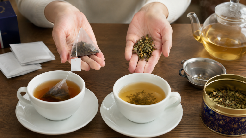 Hands comparing a pyramid tea bag and loose-leaf tea above two freshly brewed cups, with a glass teapot, tea infuser, and decorative tea tin on a wooden table.